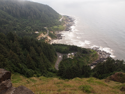 [The edge of the stone wall of the viewing platform bisects the lower left portion of the image and the curved, rocky shoreline below is visible with fog clouds hanging above all of this. In the distance is another curve of land meeting the ocean.]